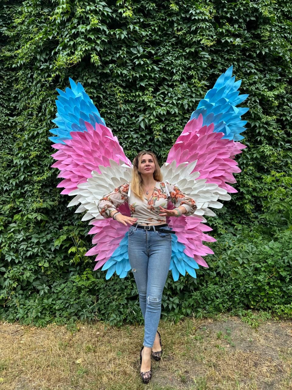 May include: A person standing in front of a wall of green foliage, wearing a floral top, blue jeans, and black heels. Large, colorful butterfly wings in shades of blue, pink, and white are behind the person.