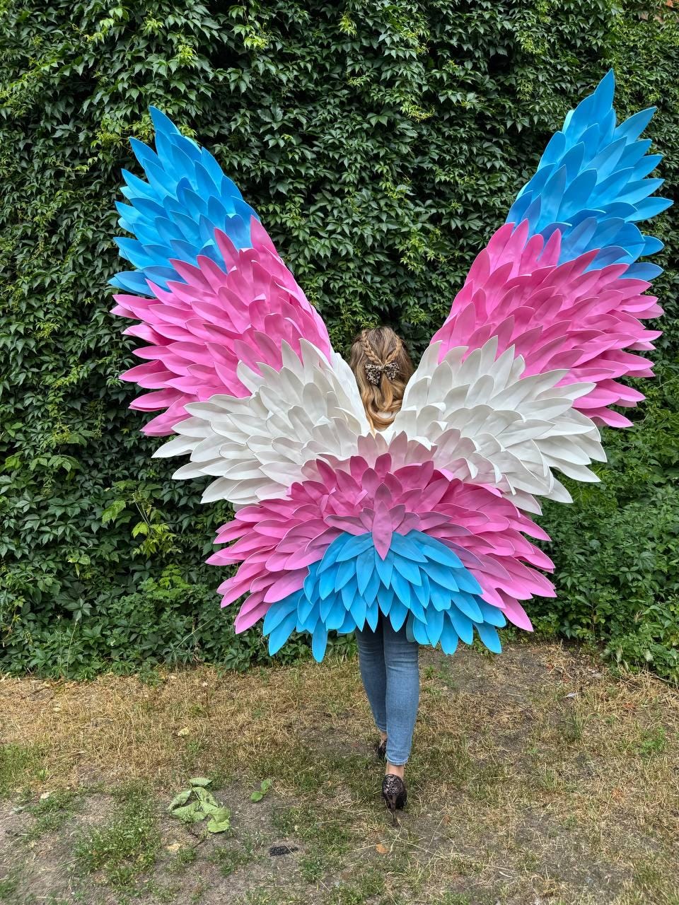 May include: Large, colorful wings in the colors of the transgender flag: light blue, pink, and white. The wings are made of layered, feather-like shapes. The person wearing the wings is standing in front of a green hedge.