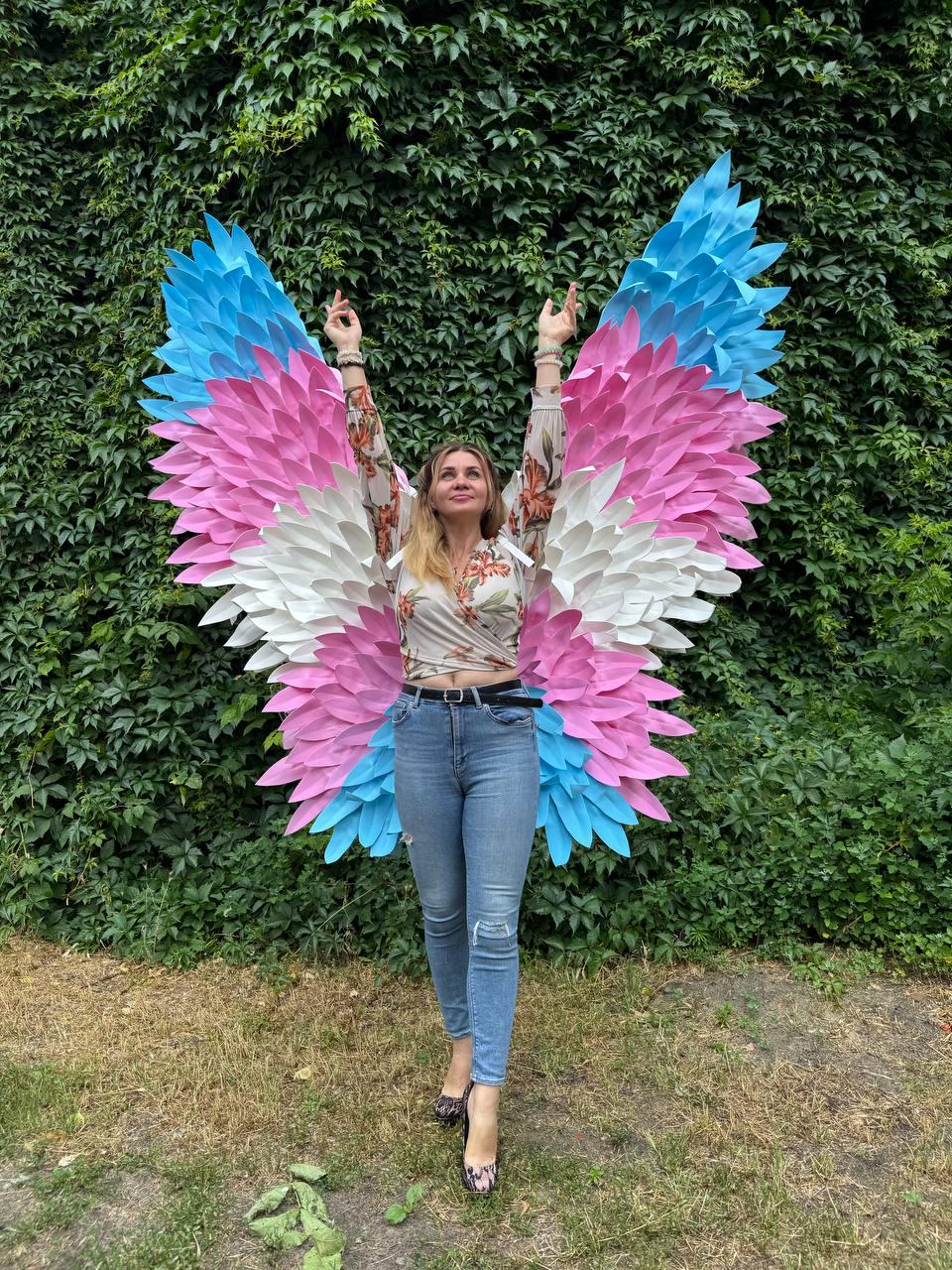 May include: Large, colorful wings in shades of blue, pink, and white, resembling feathers. The wings are worn by a person standing in front of a green leafy background. The person is wearing a floral top and blue jeans.