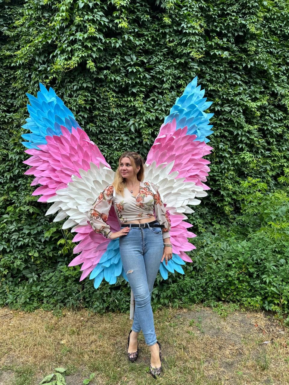 May include: Large, colorful butterfly wings in shades of pink, white, and blue. The wings are displayed against a backdrop of lush green foliage. The wings are a striking art piece.