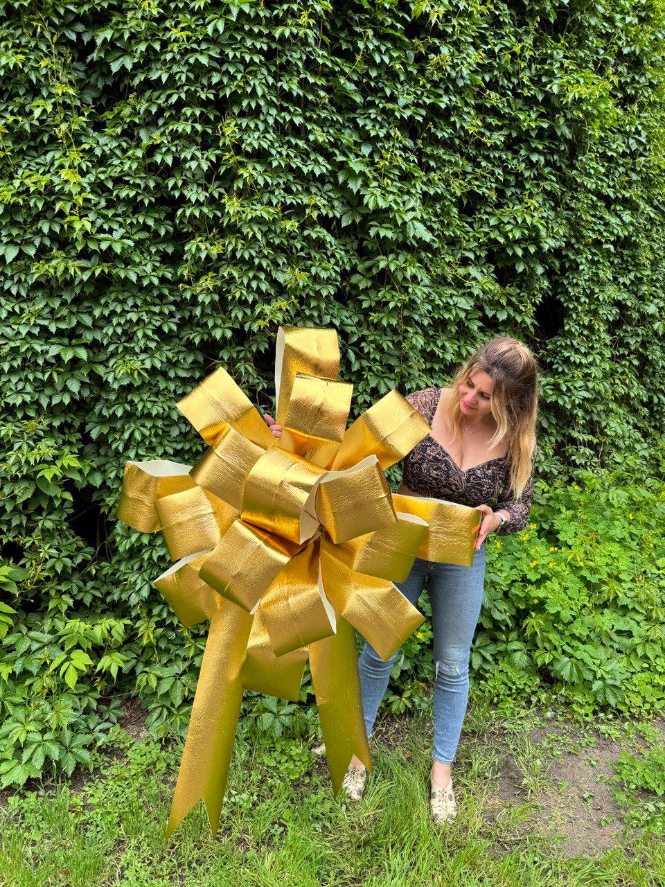 May include: A large, metallic gold decorative bow featuring multiple loops and two long, pointed tails. The bow has a textured, reflective surface and is constructed from wide ribbon material, designed for display as a prominent gift accent.