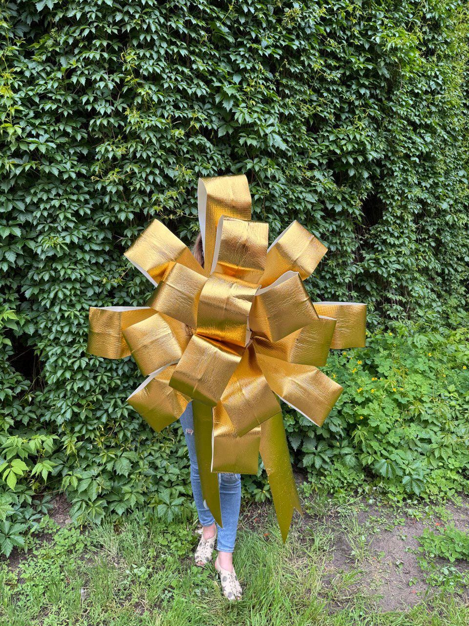 May include: A large, metallic gold decorative bow with a textured surface. The bow features multiple layered loops and two long, pointed ribbon tails extending downward, creating a full, star-like shape constructed from wide, reflective gold material.