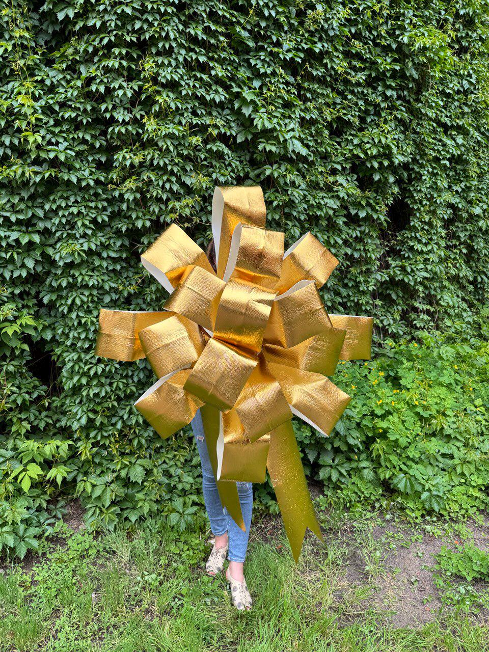 May include: A large, metallic gold decorative bow with multiple layered loops and two long, wide ribbon tails extending downward. The bow features a reflective, textured surface and is constructed from a stiff material that maintains its full, rounded shape.