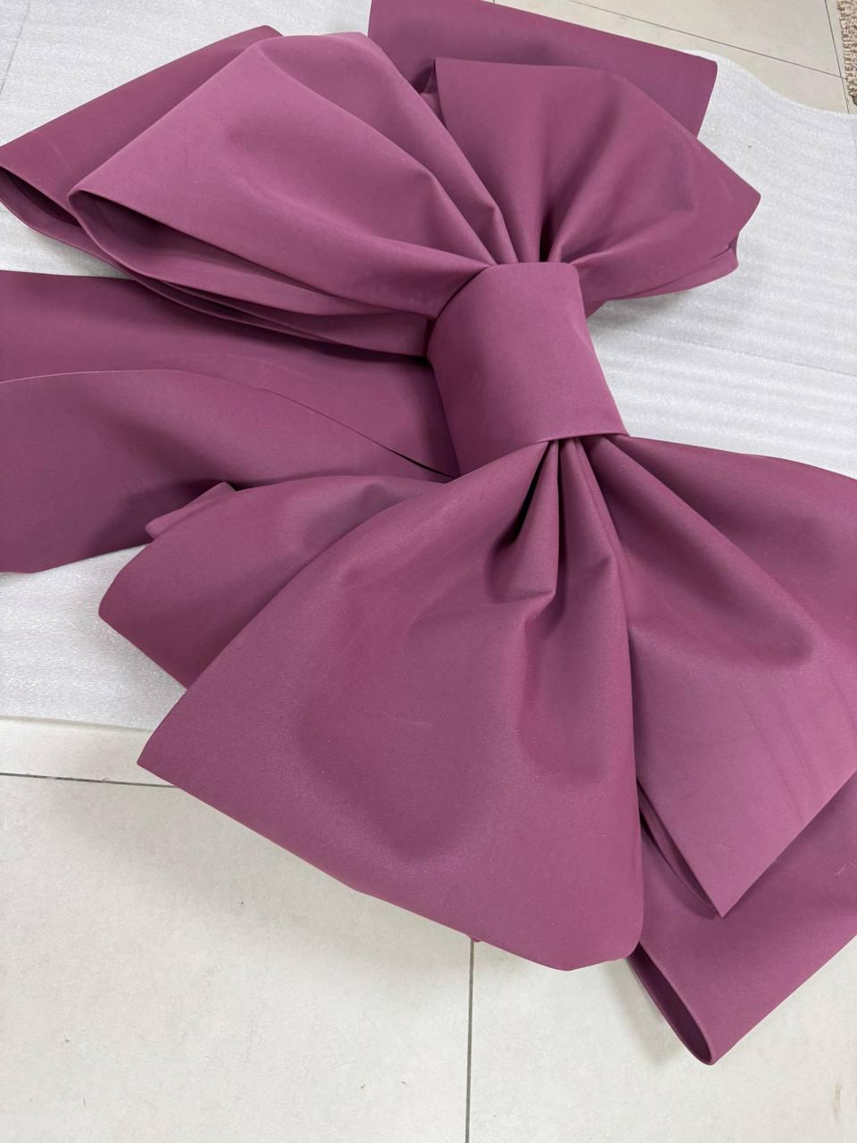 May include: A large, mauve-colored fabric bow. The bow is made of a smooth, possibly satin-like material, with multiple layers and folds creating a voluminous shape. The bow is tied in the center, with the ends cascading downwards.