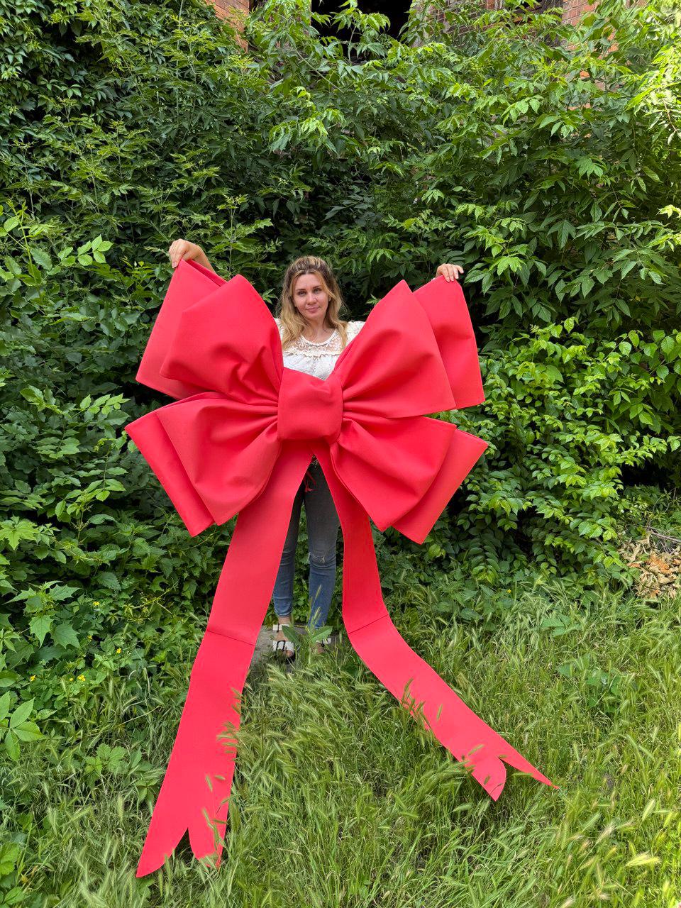 May include: A large, bright red bow with long, flowing tails. The bow is held up in front of a person standing in front of lush green foliage. The bow is made of a sturdy material and is a vibrant shade of red.