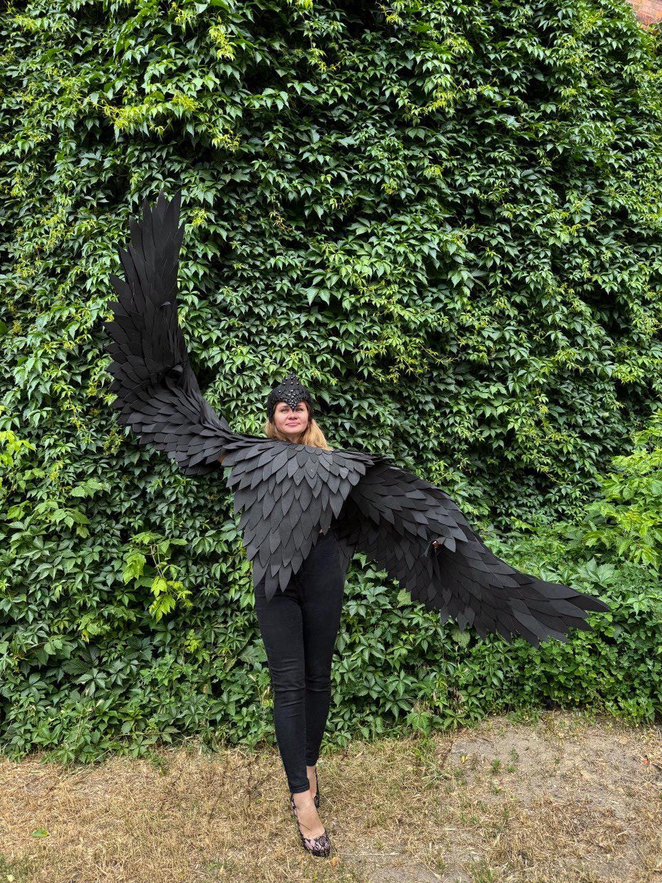 May include: A person wearing a black costume with large, feathered wings. The wings are spread wide, showcasing their impressive size and detail. The costume includes a matching headpiece and is set against a backdrop of lush green foliage.