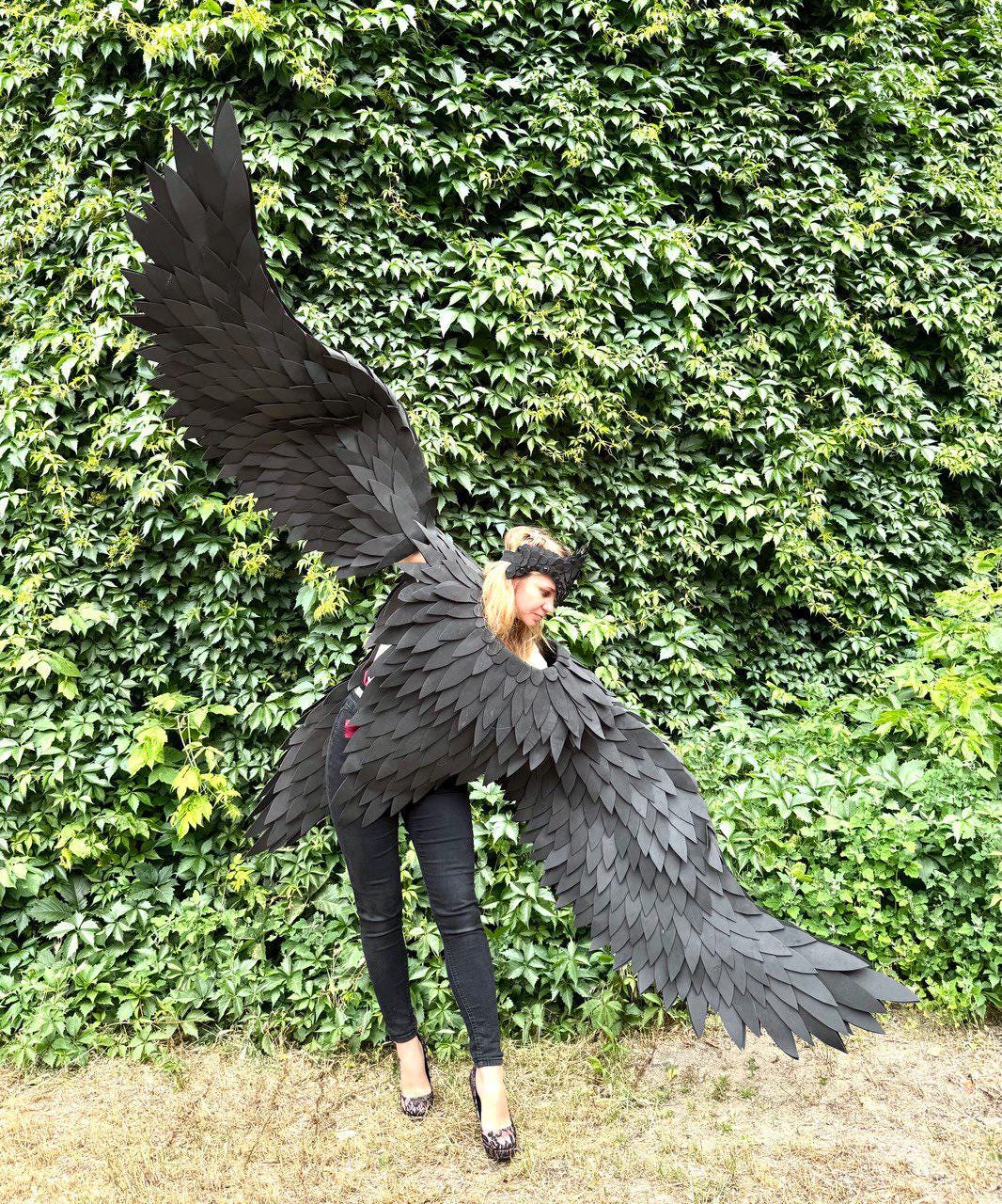 May include: A person wearing large, black, feathered costume wings, standing in front of a wall of green foliage. The wings are spread wide, showcasing their intricate design. The person is wearing black pants and heels.