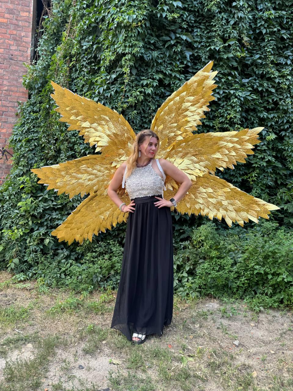 May include: A person wearing a silver and white top and a long black skirt, posing in front of a wall of green foliage. Large, golden, feathered wings are attached to the person's back, creating a striking visual contrast.