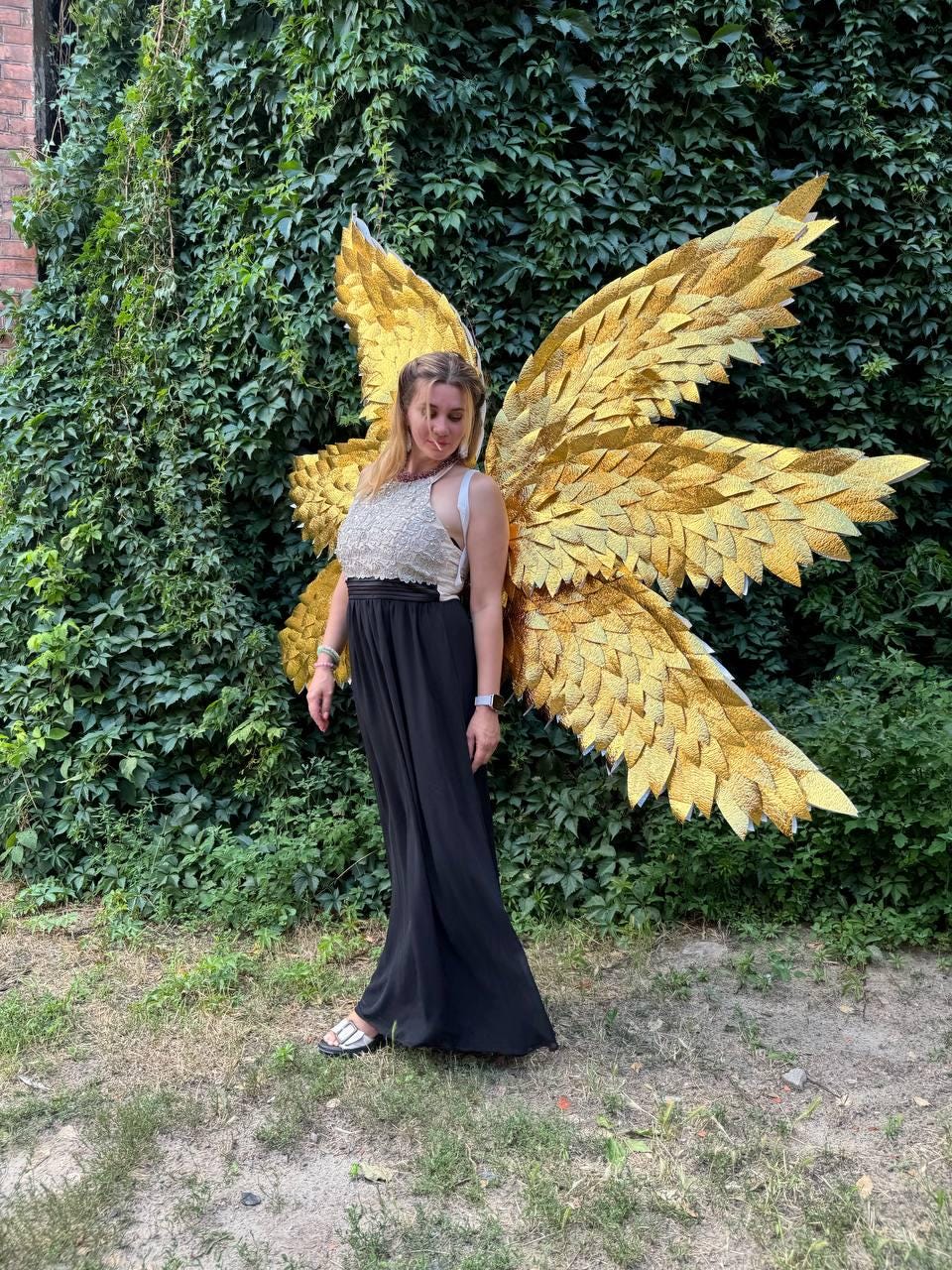May include: A person wearing large, golden, textured wings. The wings are made of many individual pieces, creating a layered effect. The person is wearing a white and gold top and a long black skirt. The background is a wall of green foliage.