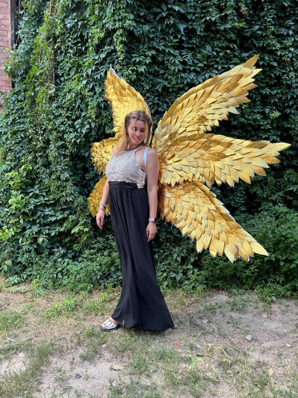 May include: A person wearing large, golden, textured wings. The wings are made of many individual pieces, creating a layered effect. The person is wearing a white and gold top and a long black skirt. The background is a wall of green foliage.