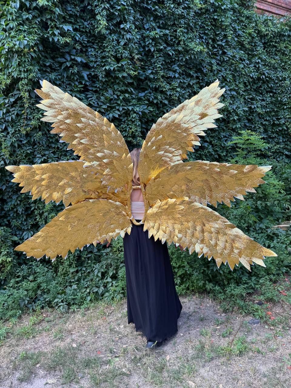 May include: Large, golden angel wings with a layered feather design. The wings are worn by a person wearing a black dress. The wings are the focal point, with a textured, metallic appearance. The background is a wall of green foliage.