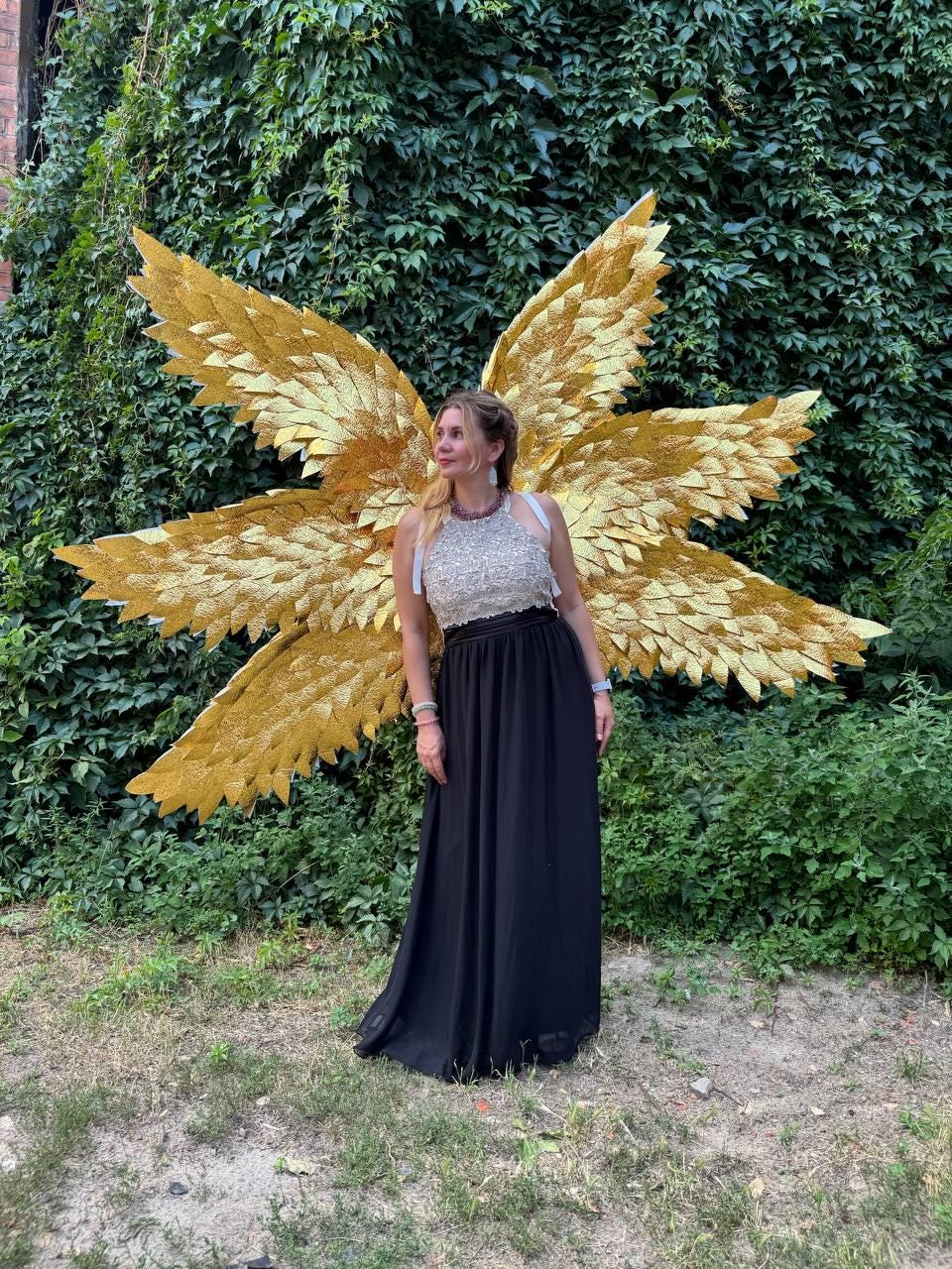 May include: A woman wearing a silver and white top and a long black skirt stands in front of a wall of green foliage. Large, golden angel wings are attached to her back, creating a striking contrast.