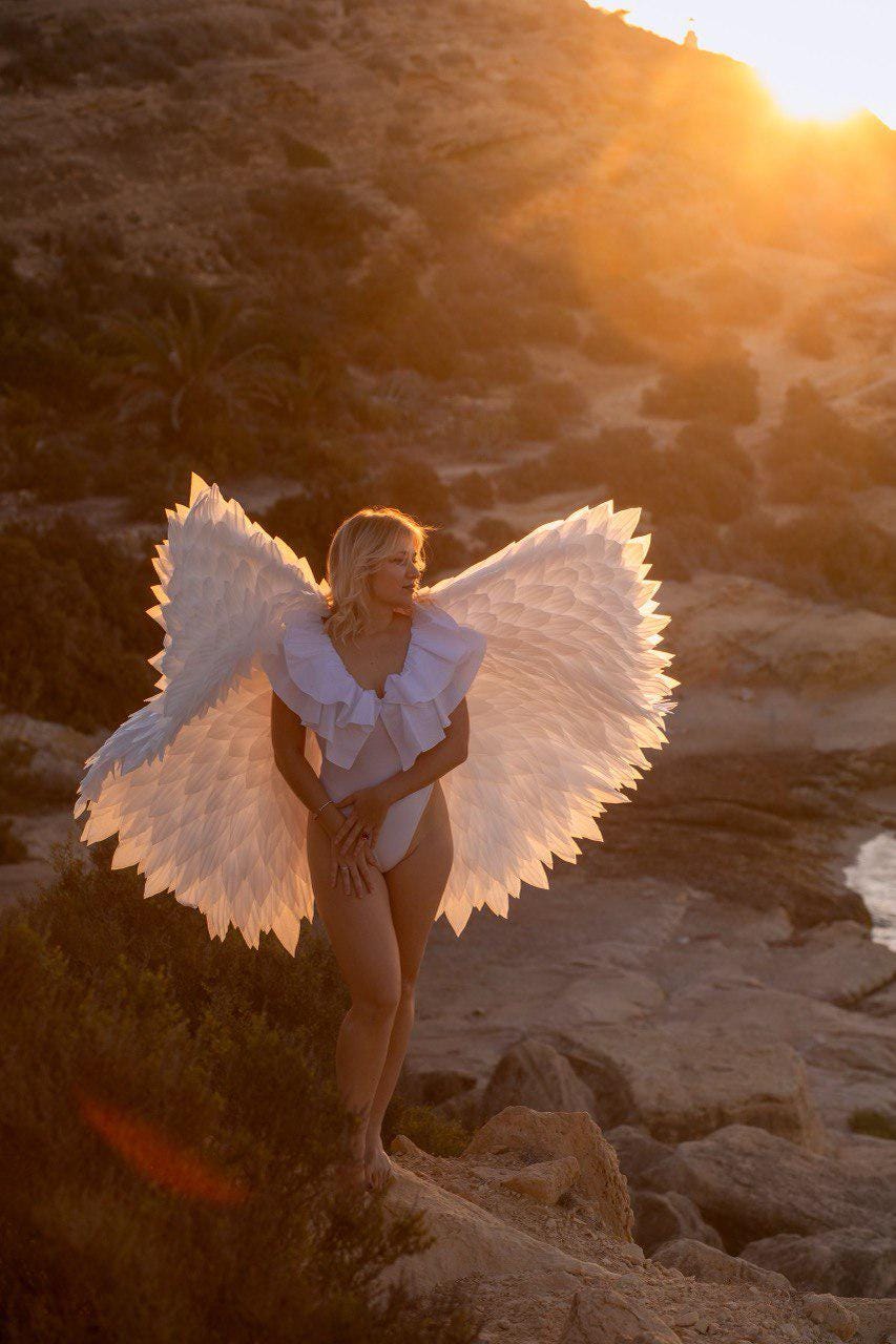 May include: A woman wearing a white bodysuit and large white feathered angel wings stands on a rocky outcrop. The wings are spread wide, catching the golden light of the setting sun. The scene evokes a sense of fantasy and ethereal beauty.
