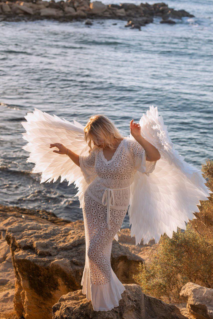 May include: A woman wearing a white, open-weave dress and large white angel wings stands on a rocky shore near the ocean. The dress has a tie at the waist and a flared hem. The wings are spread wide.