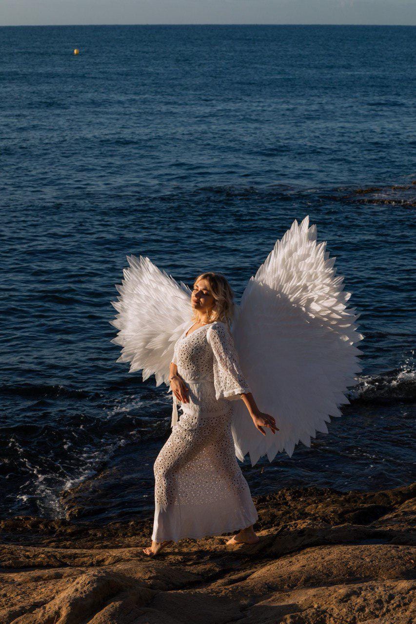 May include: A person wearing a white, long, lace dress and large white feathered angel wings stands on a rocky shore near the ocean. The person is blonde and facing the sun. The ocean is dark blue.