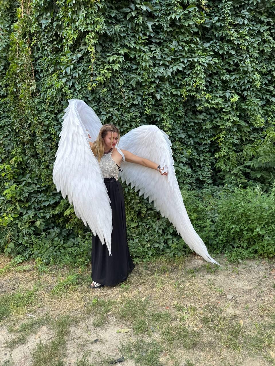 May include: A person wearing large, white angel wings, standing in front of a wall of green foliage. The wings are detailed with layered feathers. The person is wearing a black skirt and a white top.