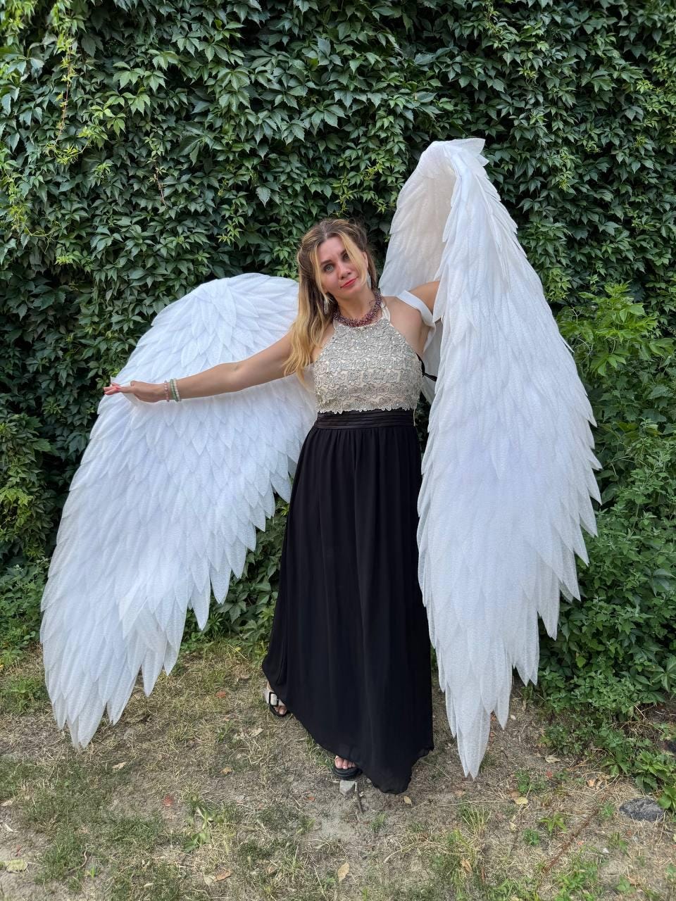 May include: A woman wearing large white feathered angel wings, a beaded halter top, and a long black skirt. The wings are spread wide against a backdrop of green foliage. The outfit is suitable for a costume or theatrical performance.