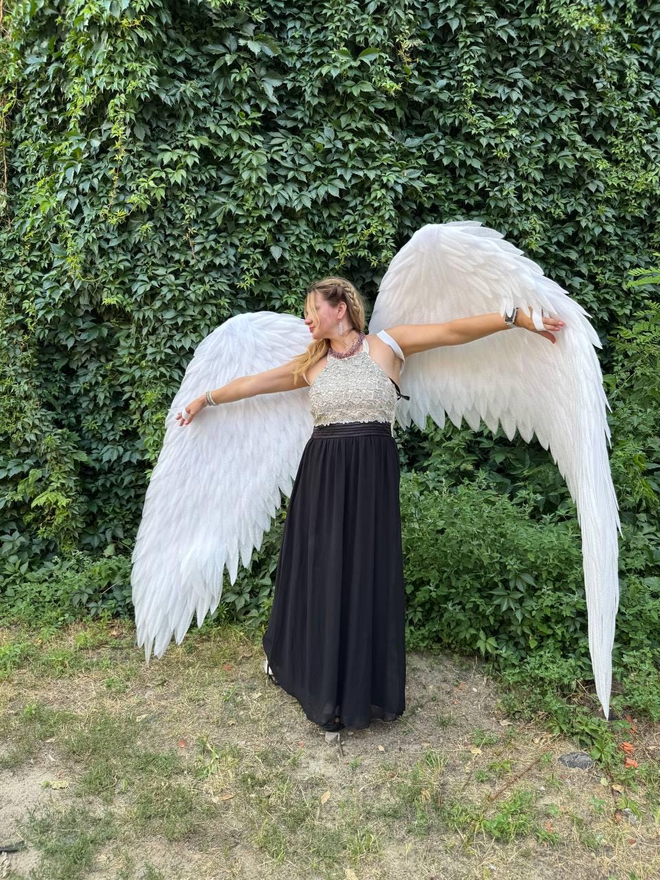May include: A person wearing large white feathered angel wings, a silver and white top, and a long black skirt, standing in front of a green leafy background. The wings are spread wide, creating a dramatic pose.