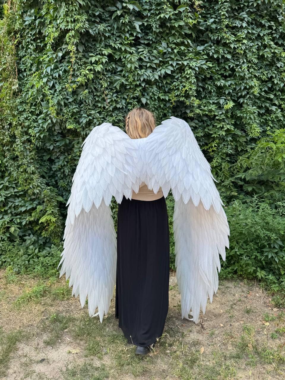 May include: A person wearing large, white angel wings, standing in front of a wall of green foliage. The wings are made of layered, textured feathers. The person is wearing a black skirt and a light-colored top.