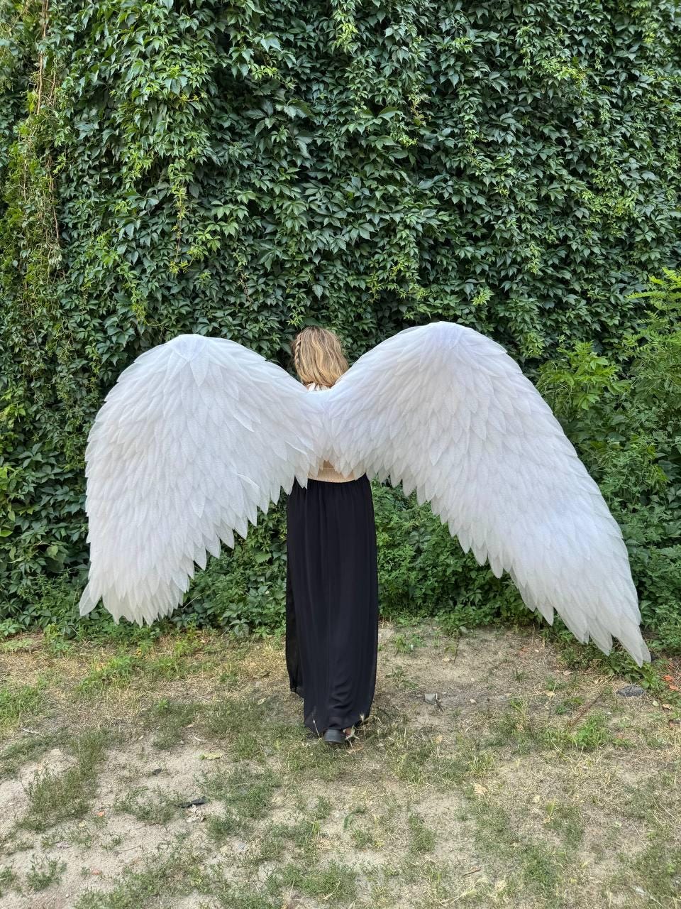 May include: Large, white feathered angel wings. The wings are spread wide, showcasing their full span. The feathers are layered, creating a textured appearance. The wings are against a backdrop of lush green foliage.