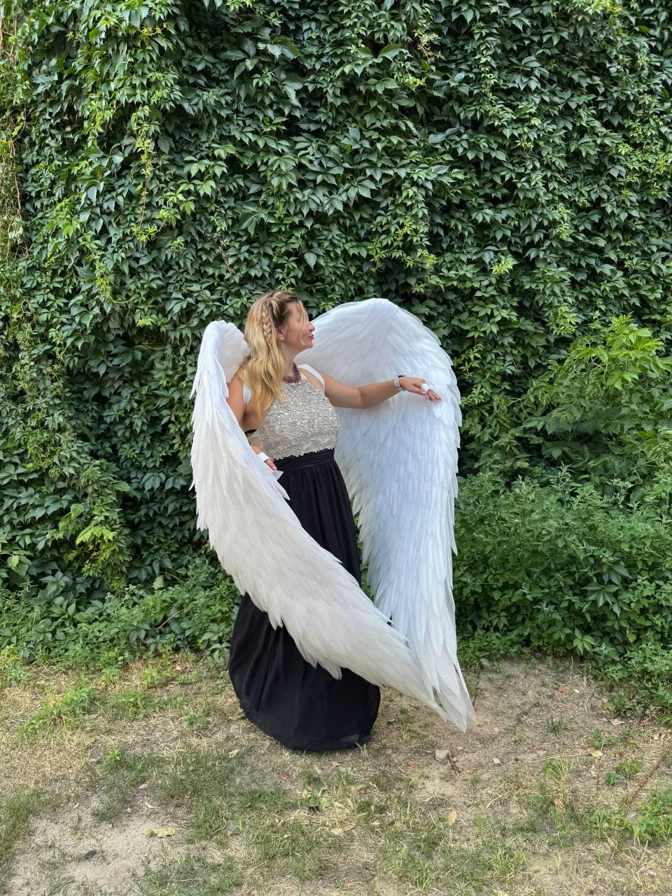 May include: A person wearing large, white angel wings, a silver top, and a black skirt, standing in front of a wall of green foliage. The wings are detailed with individual feathers, creating a dramatic effect. The outfit suggests a costume or theatrical theme.
