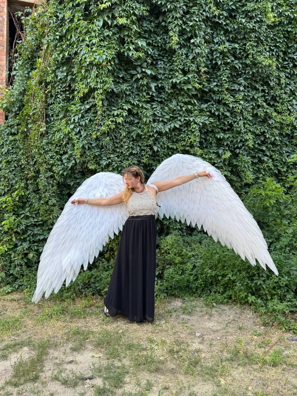 May include: A person wearing a white and gold top and a black skirt, posing with large white feathered angel wings. The wings are spread wide against a backdrop of green foliage and grass.