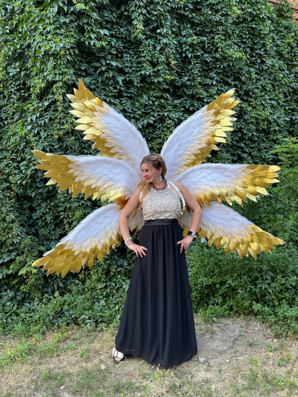 May include: A person wearing large, white and gold angel wings. The wings have white feathers with gold glitter accents. The person is wearing a black dress and a silver top. The background is a wall of green foliage.