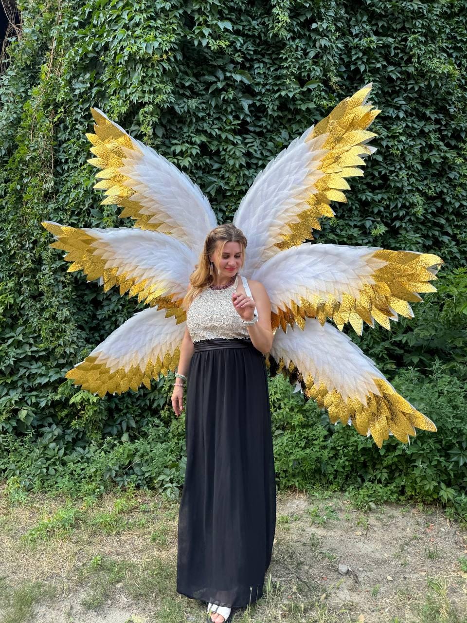 May include: A woman wearing large, ornate angel wings with white and gold feathers. The wings are the focal point, with a layered design. She is wearing a black skirt and a white top. The background is a lush green wall of foliage.