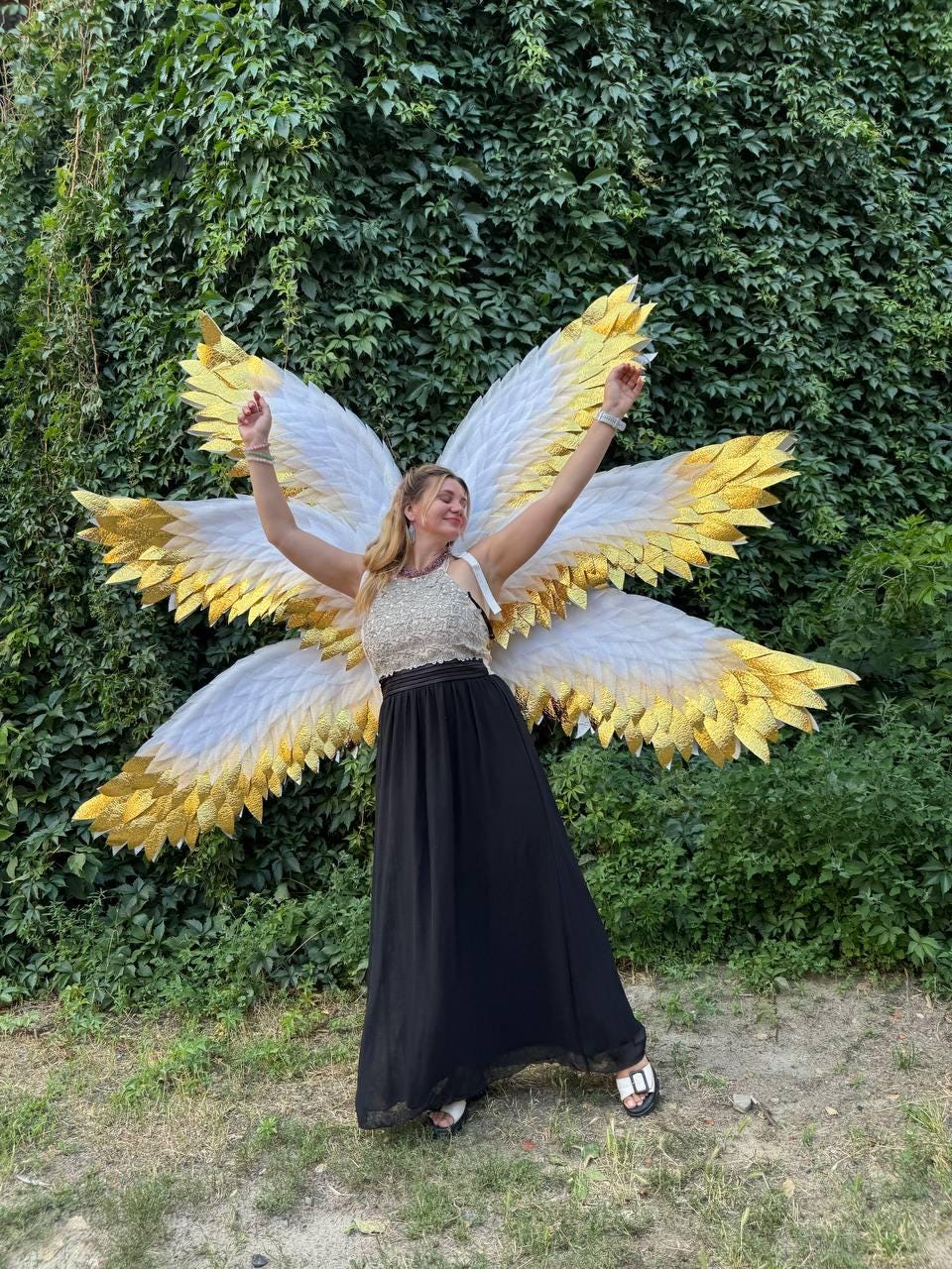 May include: A person wearing large, decorative angel wings with white and gold feathers. The wings are spread wide against a backdrop of green foliage. The person is wearing a black skirt and a sparkly top.