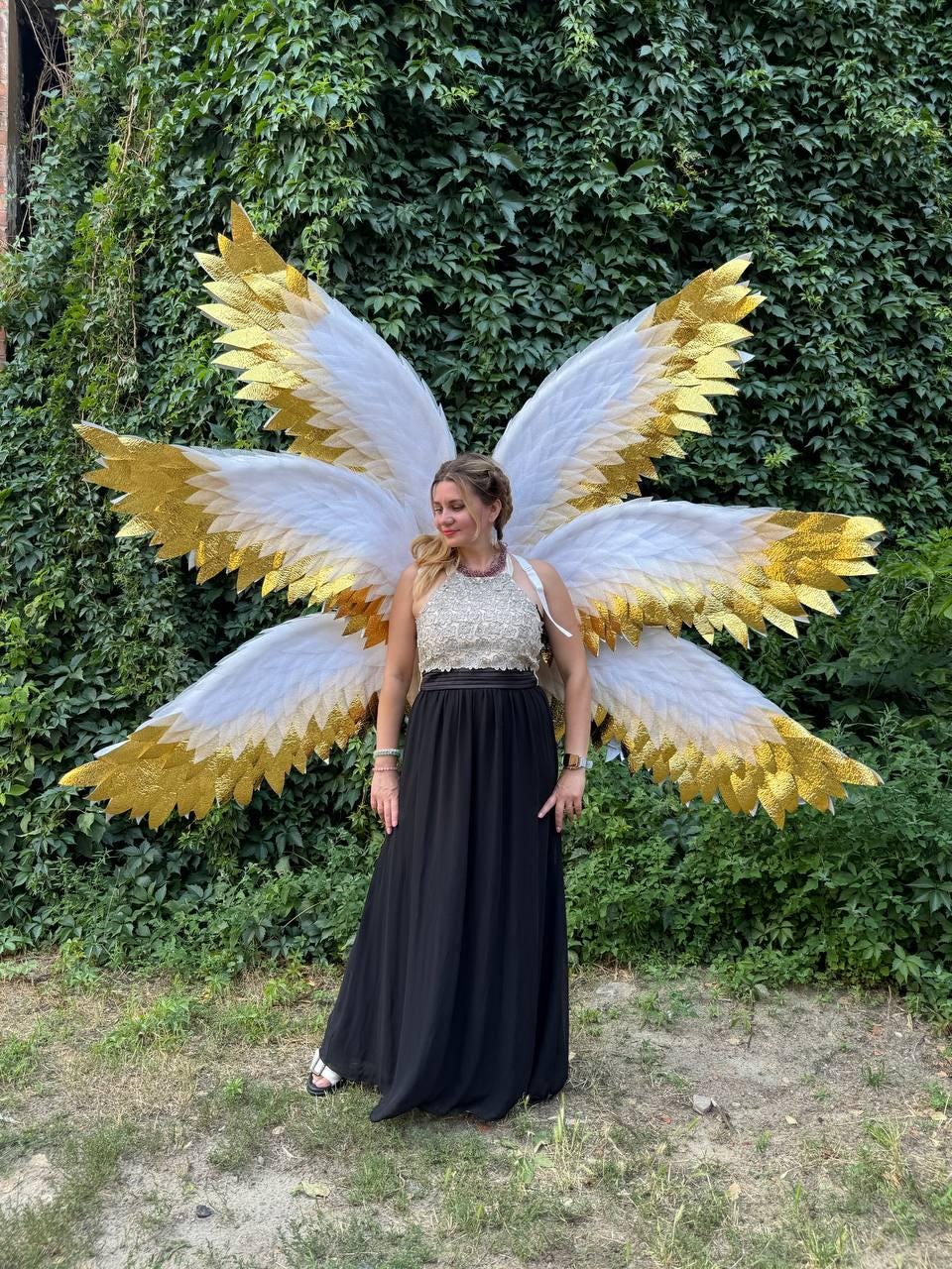 May include: A woman wearing large, elaborate angel wings with white and gold feathers. She is wearing a black floor-length skirt and a cream-colored top. The background is a wall of green foliage.