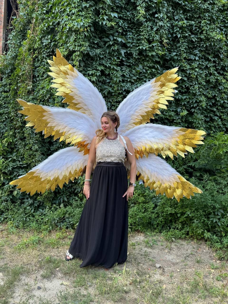 May include: A woman wearing large, decorative angel wings with white feathers and gold accents. She is wearing a black skirt and a cream-colored top. The wings are spread out against a backdrop of green foliage.