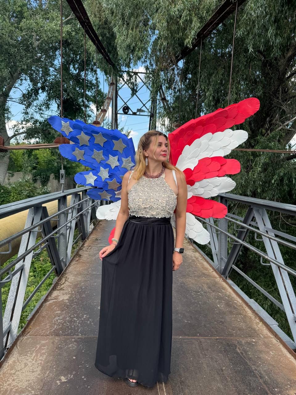 May include: A woman wearing a white and silver beaded top and a black maxi skirt stands on a bridge. Behind her are large, decorative wings in the colors of the American flag: red, white, and blue with silver stars.