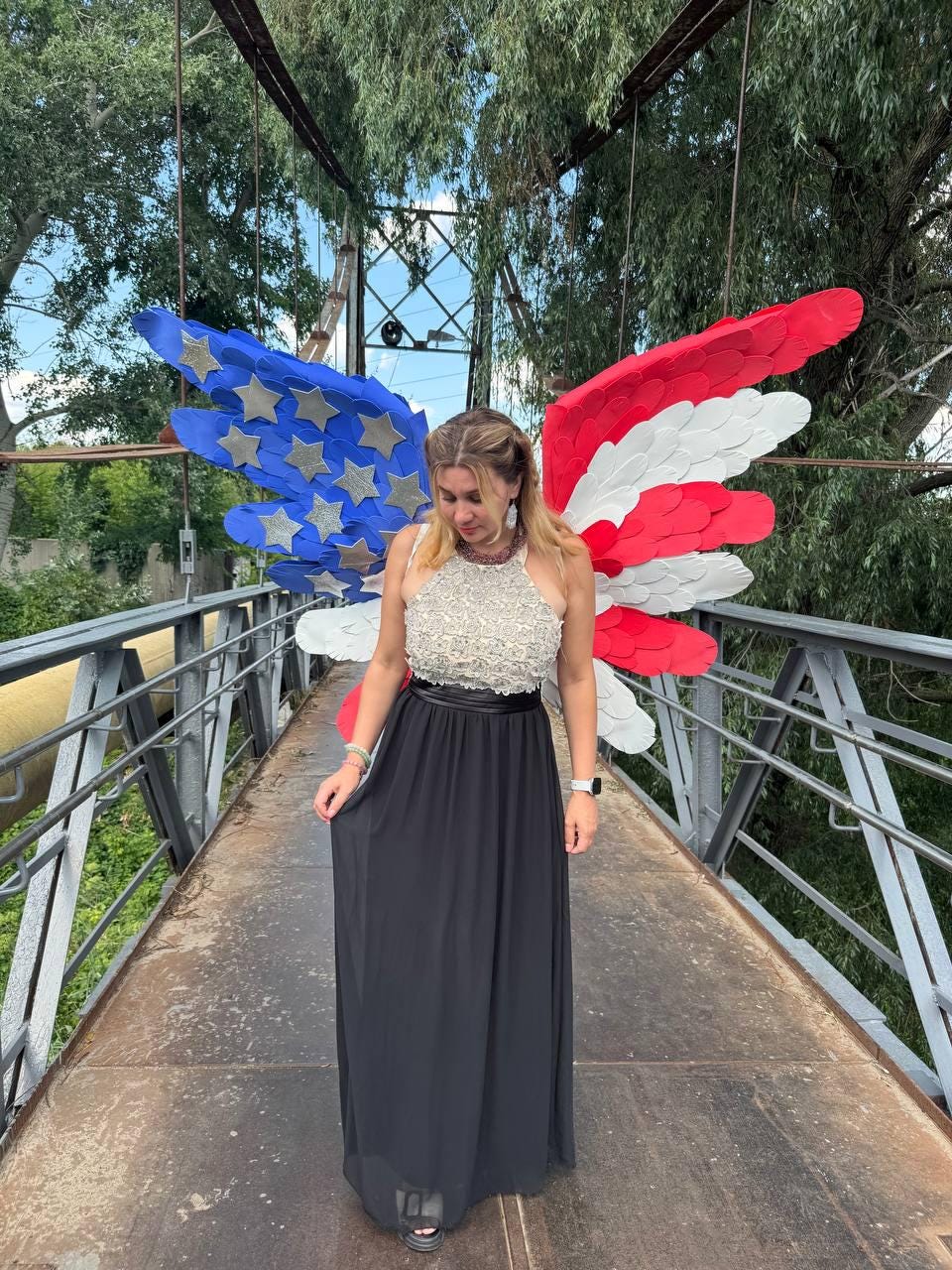 May include: A woman wearing a black floor-length dress with a beaded top stands on a bridge. Behind her are large, decorative wings in the colors of the American flag: red, white, and blue. The bridge has metal railings and a yellow pipe.