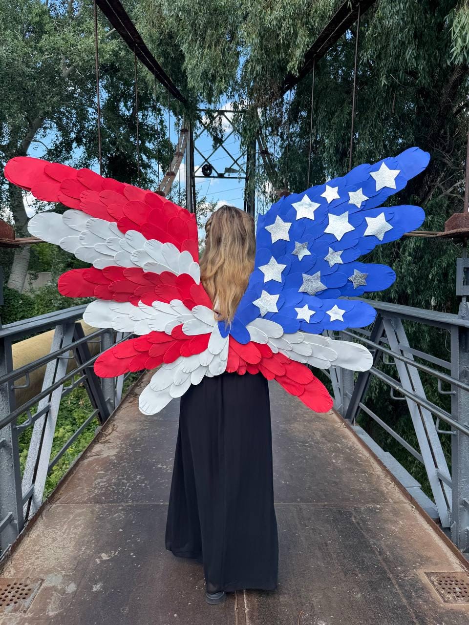 May include: A person wearing patriotic wings, designed with red, white, and blue colors. The wings are made to resemble the American flag, with red and white stripes on one side and blue with white stars on the other. The person is wearing a long black dress.