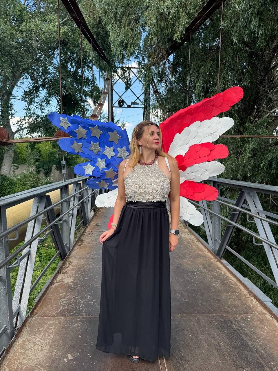 May include: A woman stands on a bridge, wearing a formal black skirt and a beaded top. Behind her are large, colorful wings in the style of the American flag, with blue, white, and red sections. The bridge has metal railings and a metal surface.