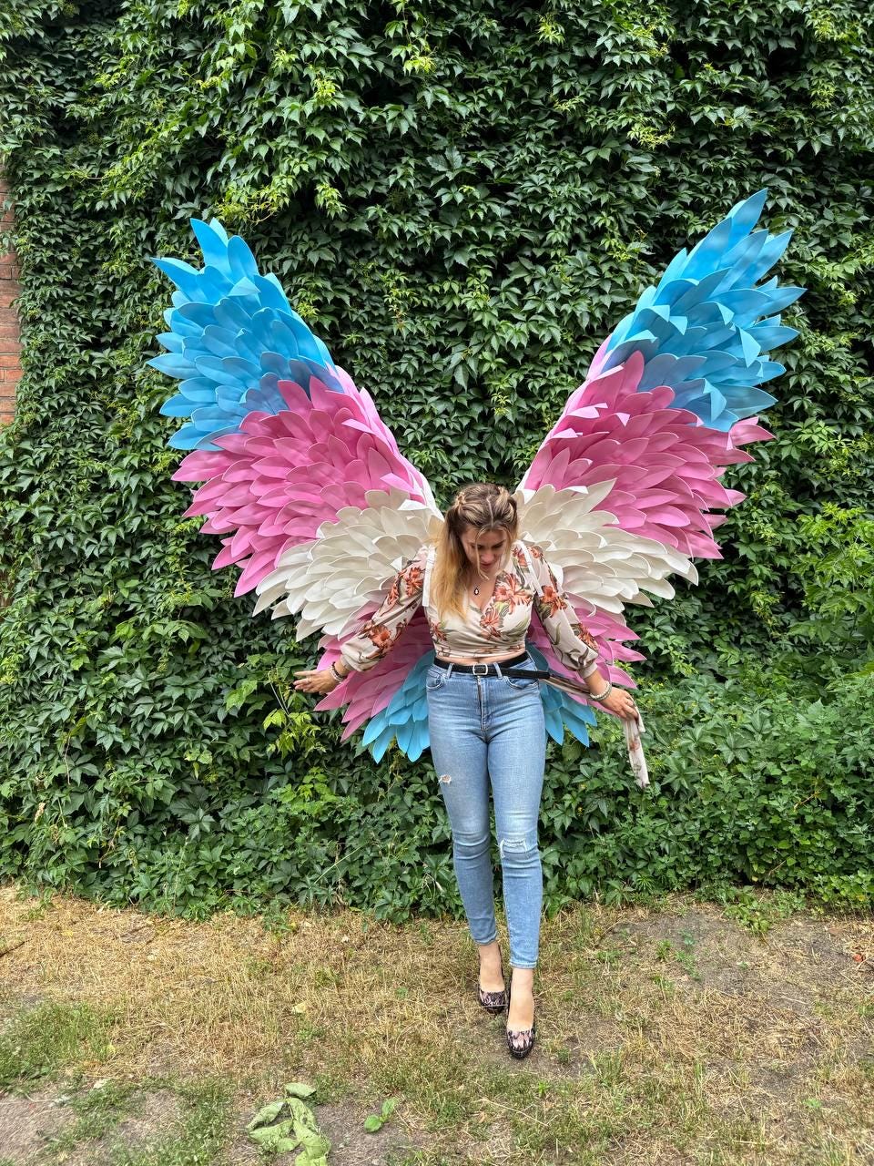 May include: Large, colorful angel wings with blue, pink, and white feathers. The wings are displayed against a backdrop of lush green foliage. The wings are a unique costume piece.