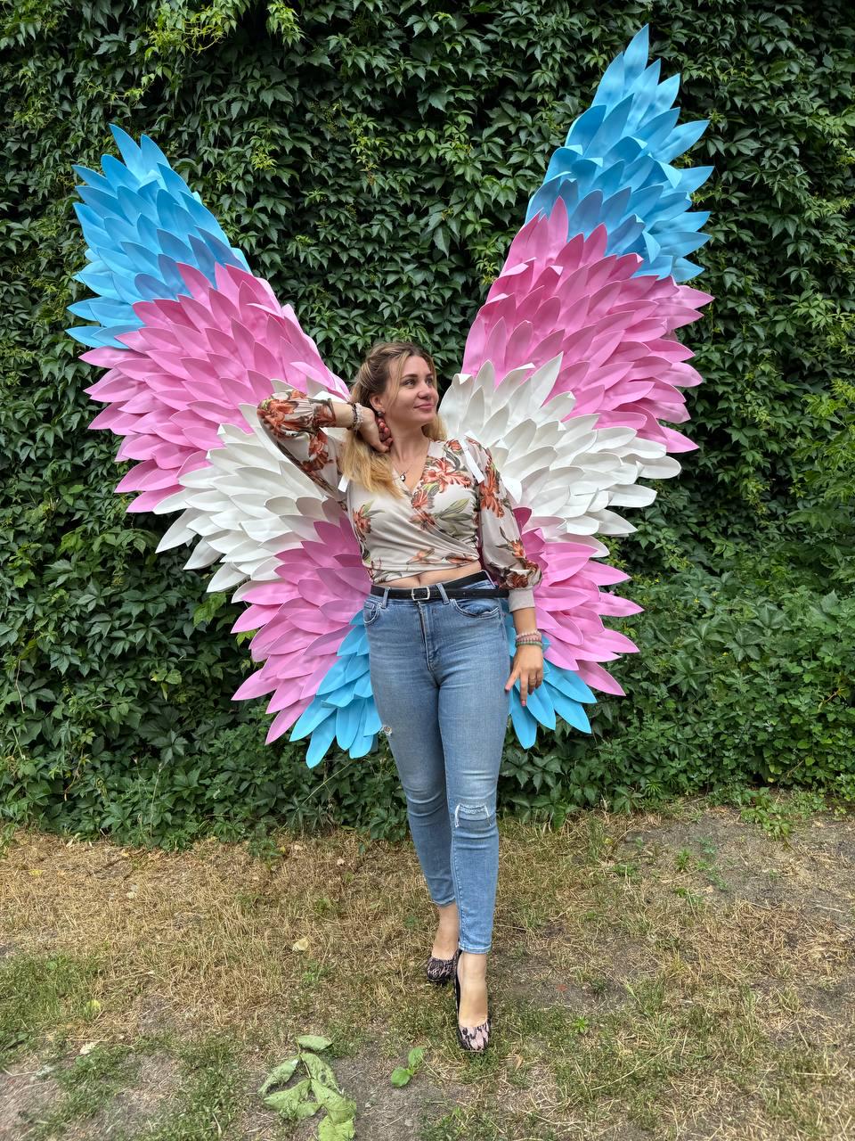 May include: Large, colorful angel wings in shades of blue, pink, and white. The wings are displayed against a backdrop of green foliage. The person in the image is wearing a floral top, blue jeans, and patterned heels.