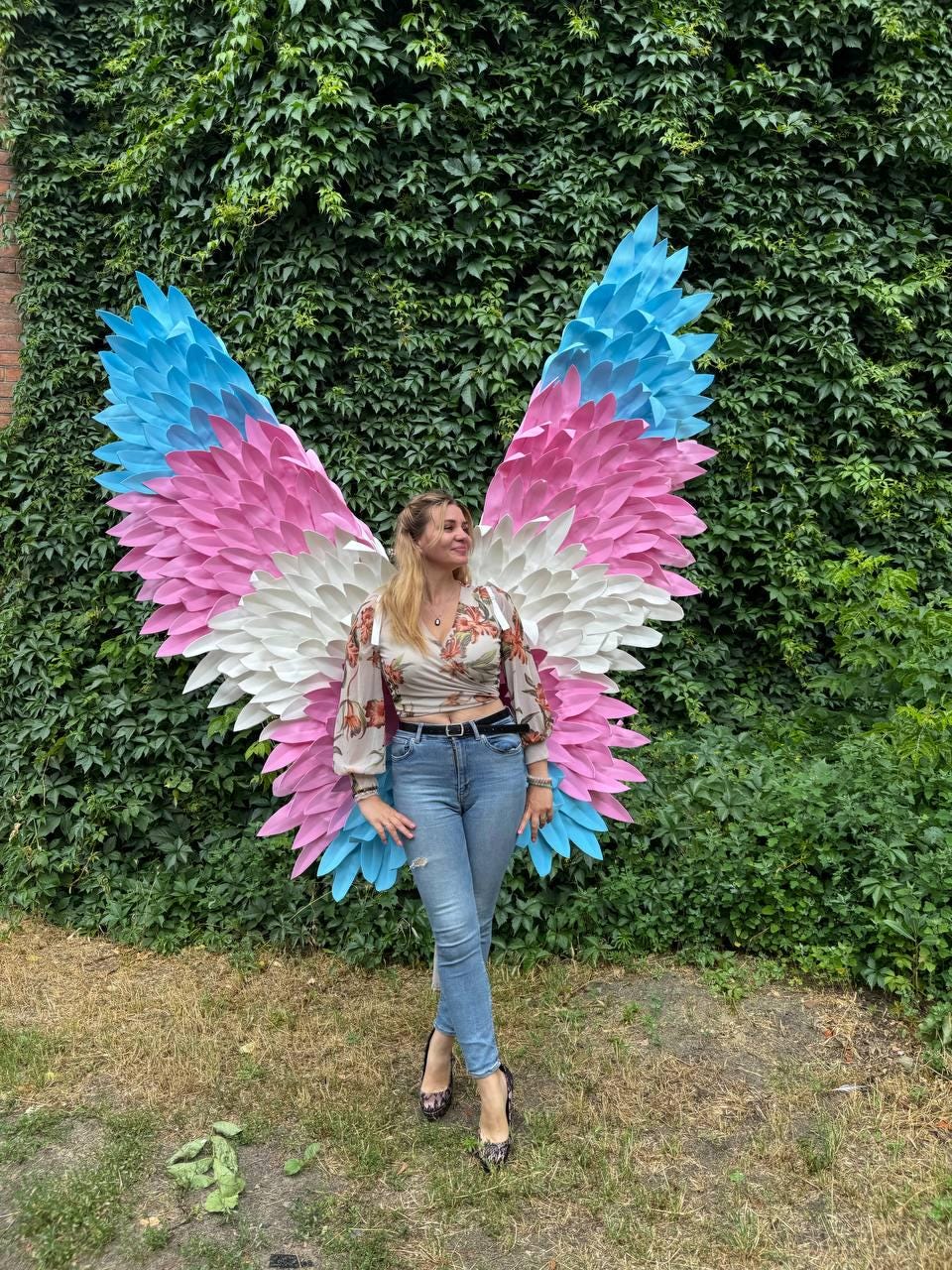May include: Large, colorful butterfly wings with blue, pink, and white feathers are displayed against a backdrop of green foliage. The wings are a vibrant art installation, perfect for photo opportunities.
