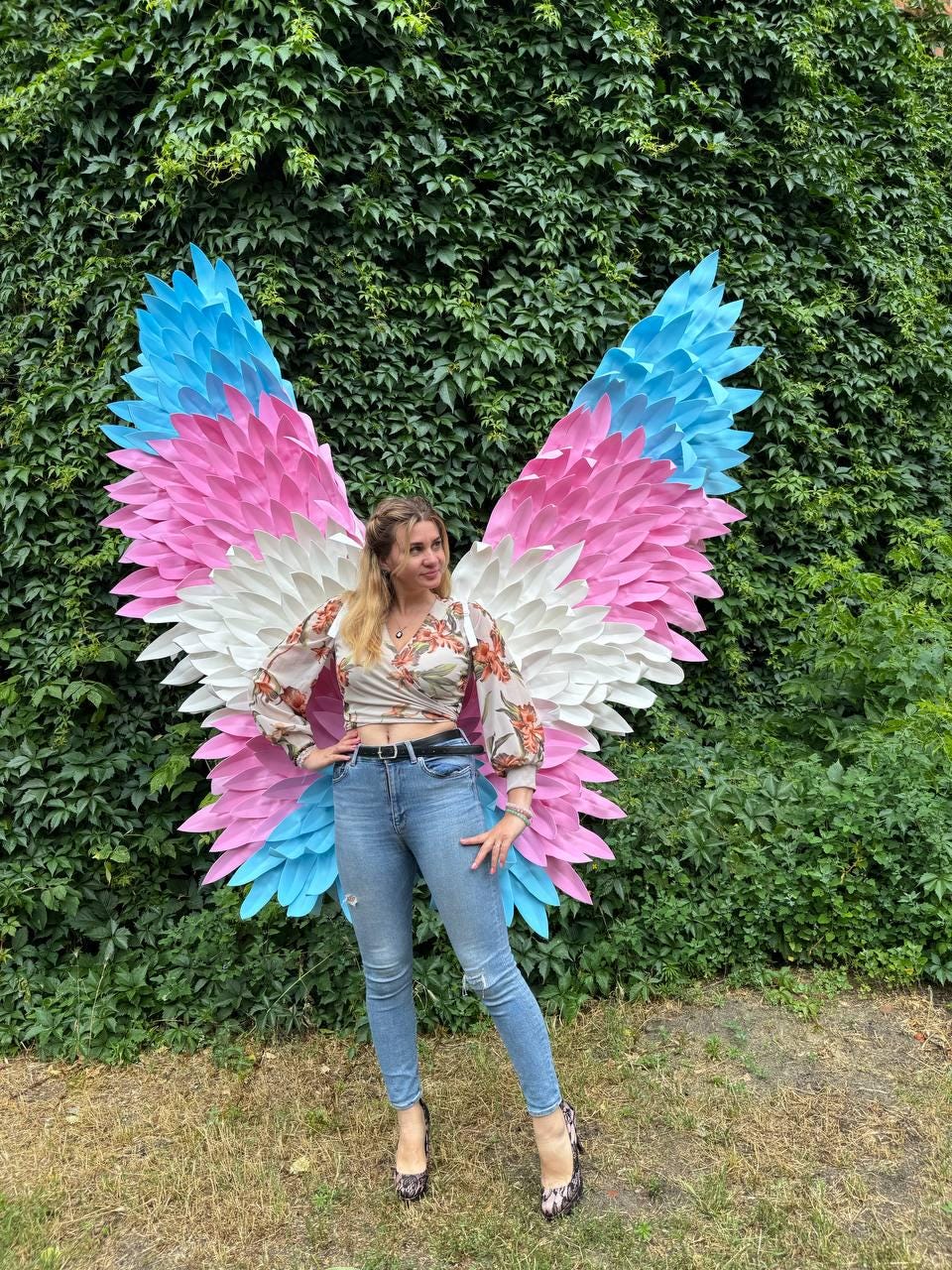 May include: Large, colorful wings in the colors of the transgender flag: light blue, pink, and white. The wings are made of layered, textured material. The wings are displayed in front of a wall of green foliage.