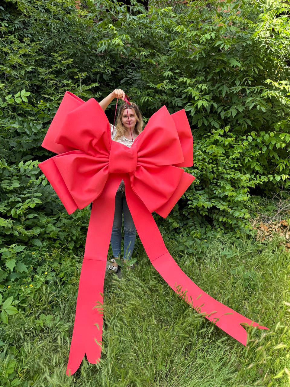 May include: A large, bright red bow with long streamers. The bow is held up against a backdrop of green foliage. The bow is made of a sturdy material and is designed for decorative purposes.