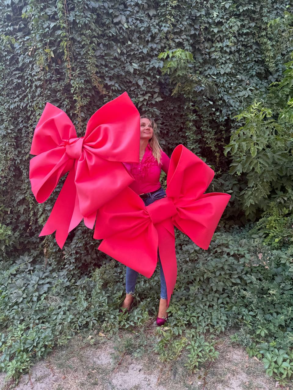 May include: Two large, vibrant red bows. The bows are the focal point, with a woman in a pink top and jeans visible behind them. The background is a lush green wall of foliage. The bows have long, flowing tails.