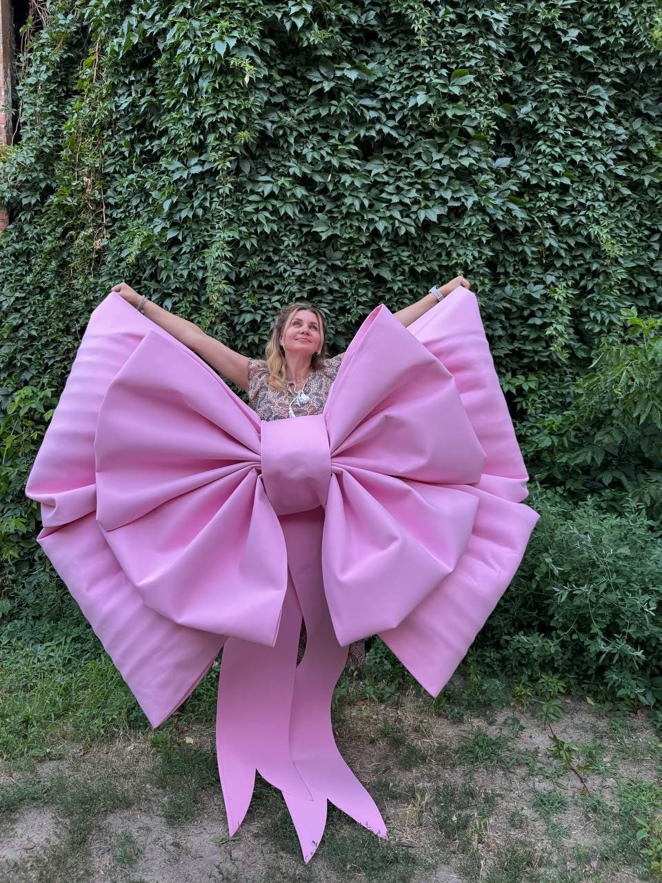 May include: A large, pink bow with long, flowing ribbons. The bow is made of a smooth material and is held up in front of a green leafy background. The bow is a vibrant shade of pink and appears to be a decorative item.