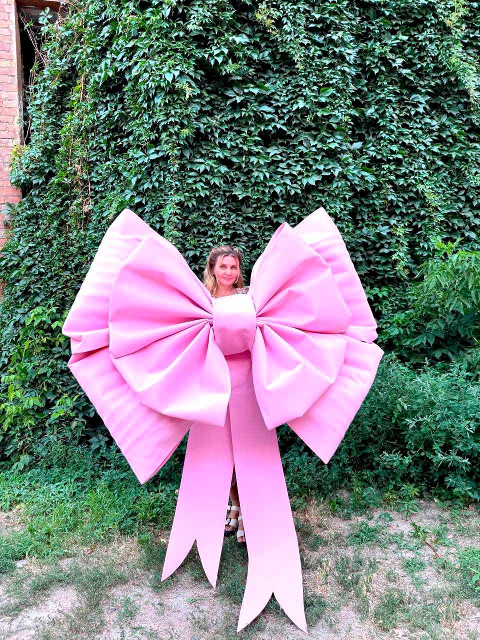 May include: A large, pink bow with long, flowing ribbons. The bow is made of a smooth, satin-like material and is positioned in front of a lush green wall of foliage. The bow's size suggests it could be used for decoration or as a prop.