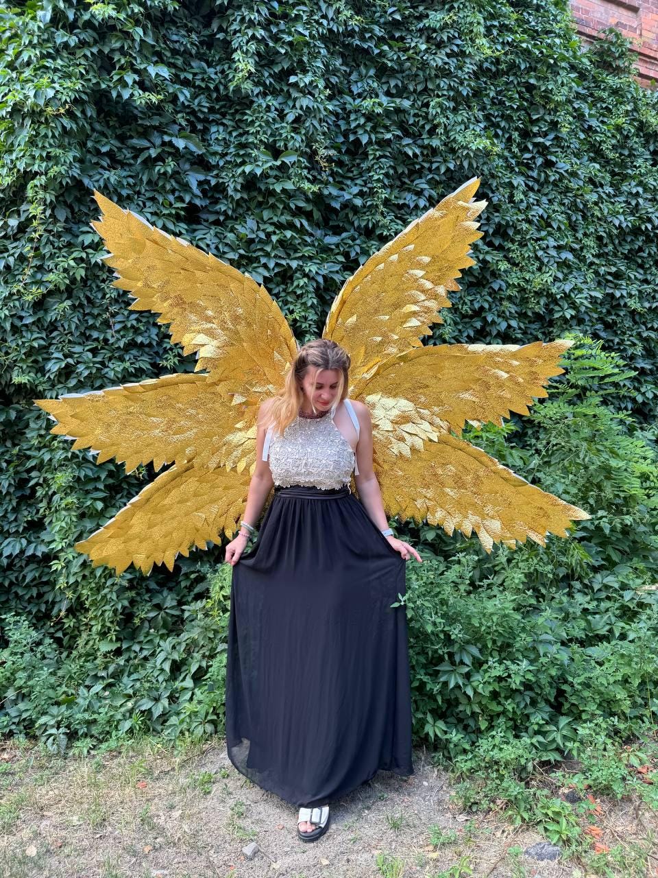 May include: A person wearing a gold sequined top and a long black skirt, with large golden angel wings. The wings are detailed and textured, set against a backdrop of green foliage. The outfit is suitable for a costume or special event.