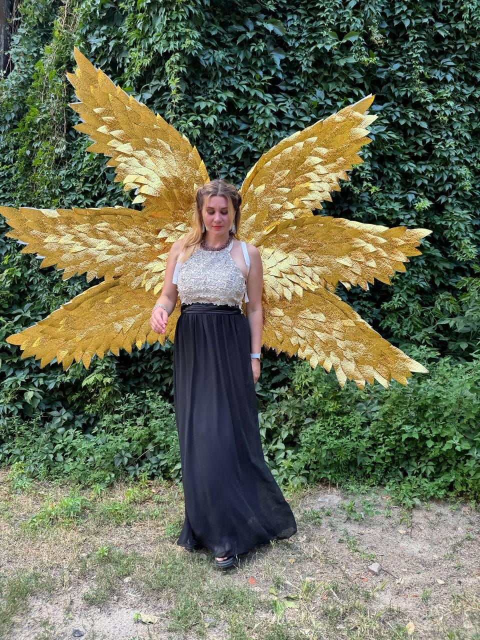 May include: A person wearing a gold-colored angel wing costume with a black skirt and a silver top. The wings are large and detailed, with a layered feather design. The background is a lush green wall of foliage.