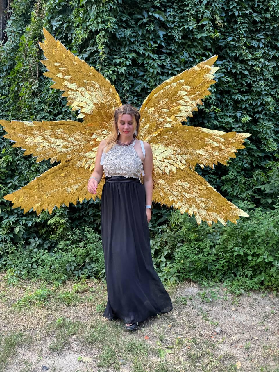 May include: A person wearing a gold-colored angel wing costume with a black skirt and a silver top. The wings are large and detailed, with a layered feather design. The background is a lush green wall of foliage.
