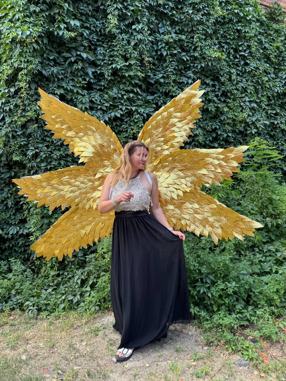 May include: A person wearing a gold glitter angel wing costume with a black maxi skirt and a silver top. The wings are large and detailed, with a layered design. The background is a lush green wall of foliage.