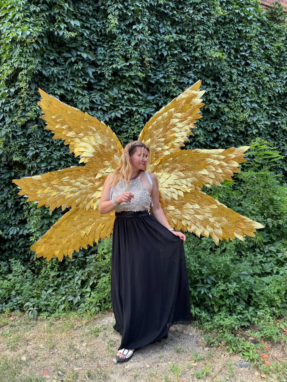 May include: A person wearing a gold glitter angel wing costume with a black maxi skirt and a silver top. The wings are large and detailed, with a layered design. The background is a lush green wall of foliage.