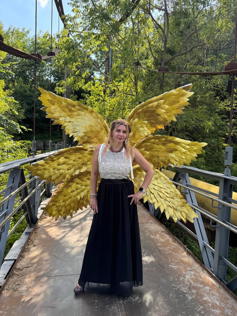 May include: A woman wearing a white and silver embellished top and a long black skirt stands on a bridge. Large, golden, feathered wings are behind her, creating a striking visual. The bridge has metal railings and is surrounded by lush green trees.