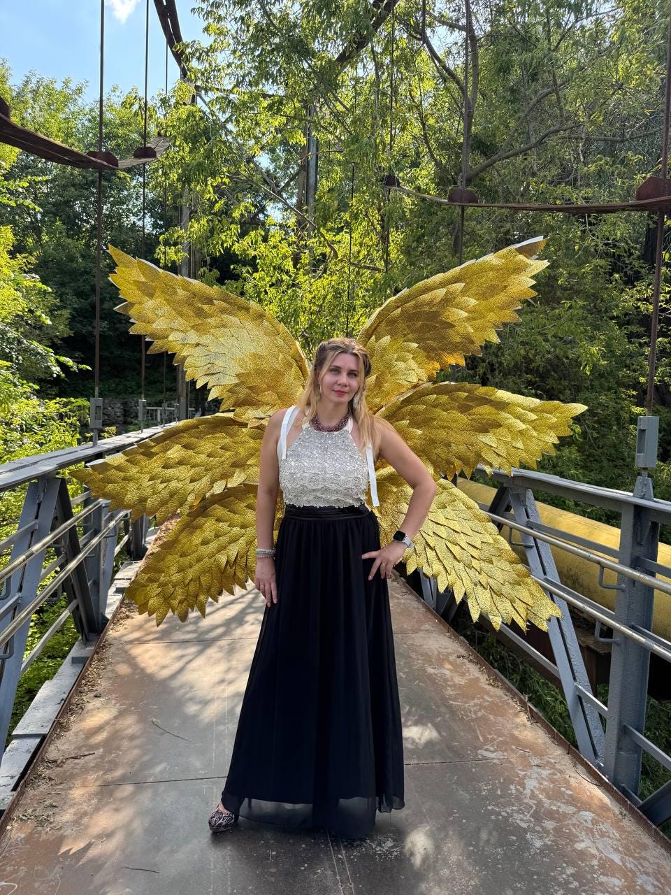 May include: A woman wearing a white and silver embellished top and a long black skirt stands on a bridge. Large, golden, feathered wings are behind her, creating a striking visual. The bridge has metal railings and is surrounded by lush green trees.